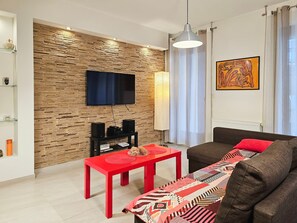 Un salón moderno con una pared de diseño que imita la piedra, un televisor montado, una mesa roja, un sofá marrón y una manta de colores.