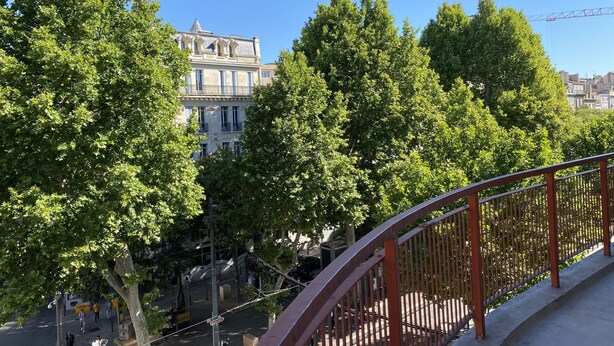 Un balcon doté d’une balustrade en métal, donnant sur une rue bordée d’arbres et sur un bâtiment coiffé d’un dôme, visible au loin.