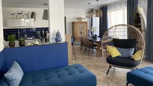 Un salon moderne avec un canapé bleu, un fauteuil en osier agrémenté de coussins, une table à manger et un coin cuisine avec des carreaux bleus et des objets décoratifs.