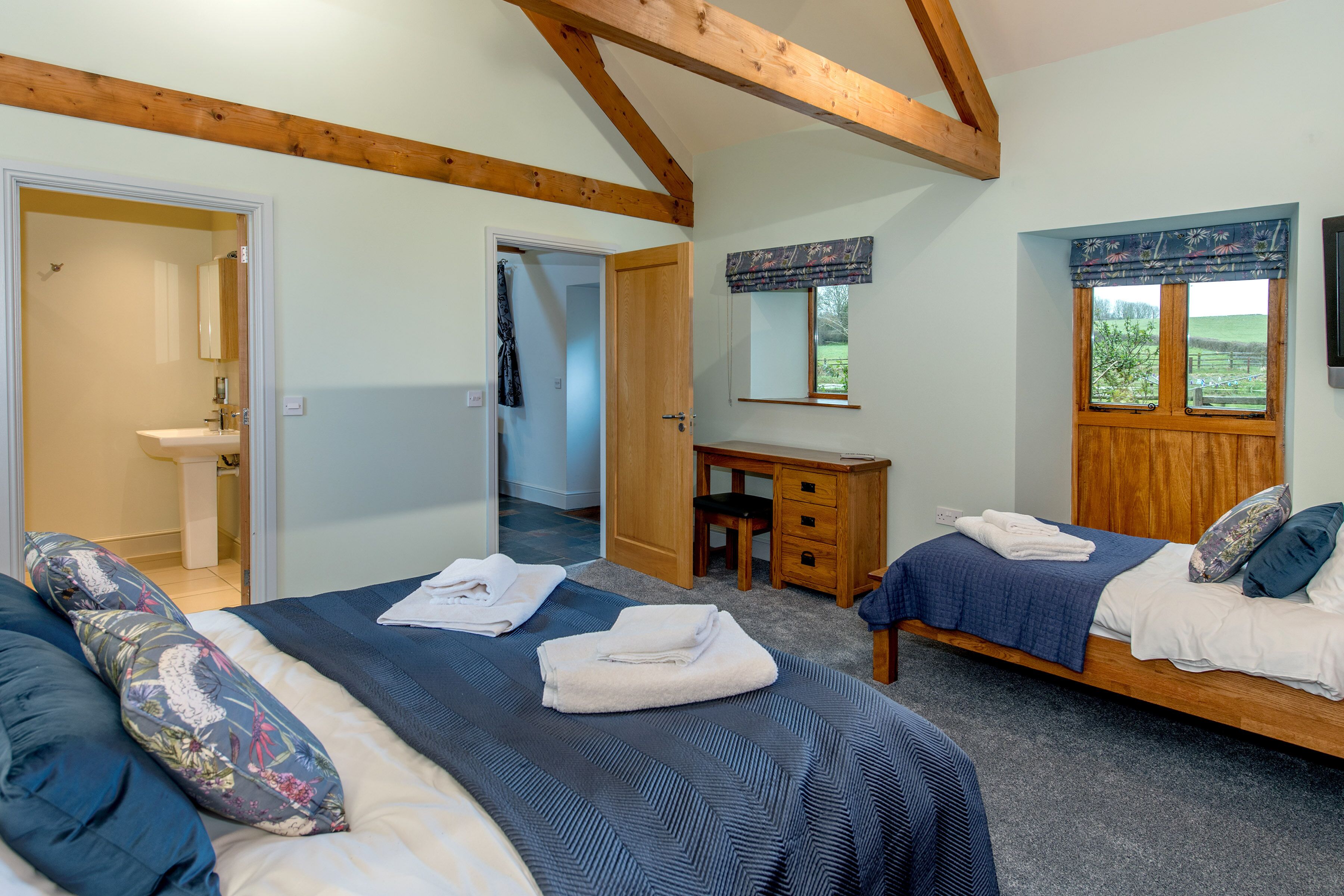Une chambre avec deux lits, un bureau en bois, un miroir et une fenêtre donnant sur un paysage verdoyant.
