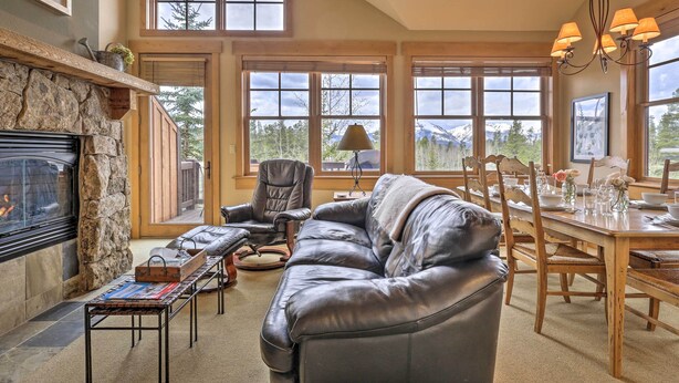 A cozy living room with a stone fireplace, leather sofa, dining table, and large windows offering mountain views. - 3 Mi to Keystone Resort: Condo w/ Mountain View (Keystone)