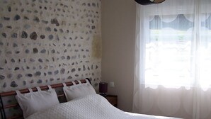 A bedroom with a stone-patterned wall, a bed with white bedding, a wooden headboard, a bedside table with a lamp, and a window with sheer curtains. - Authentic cottage in the countryside near Pau (Cescau)