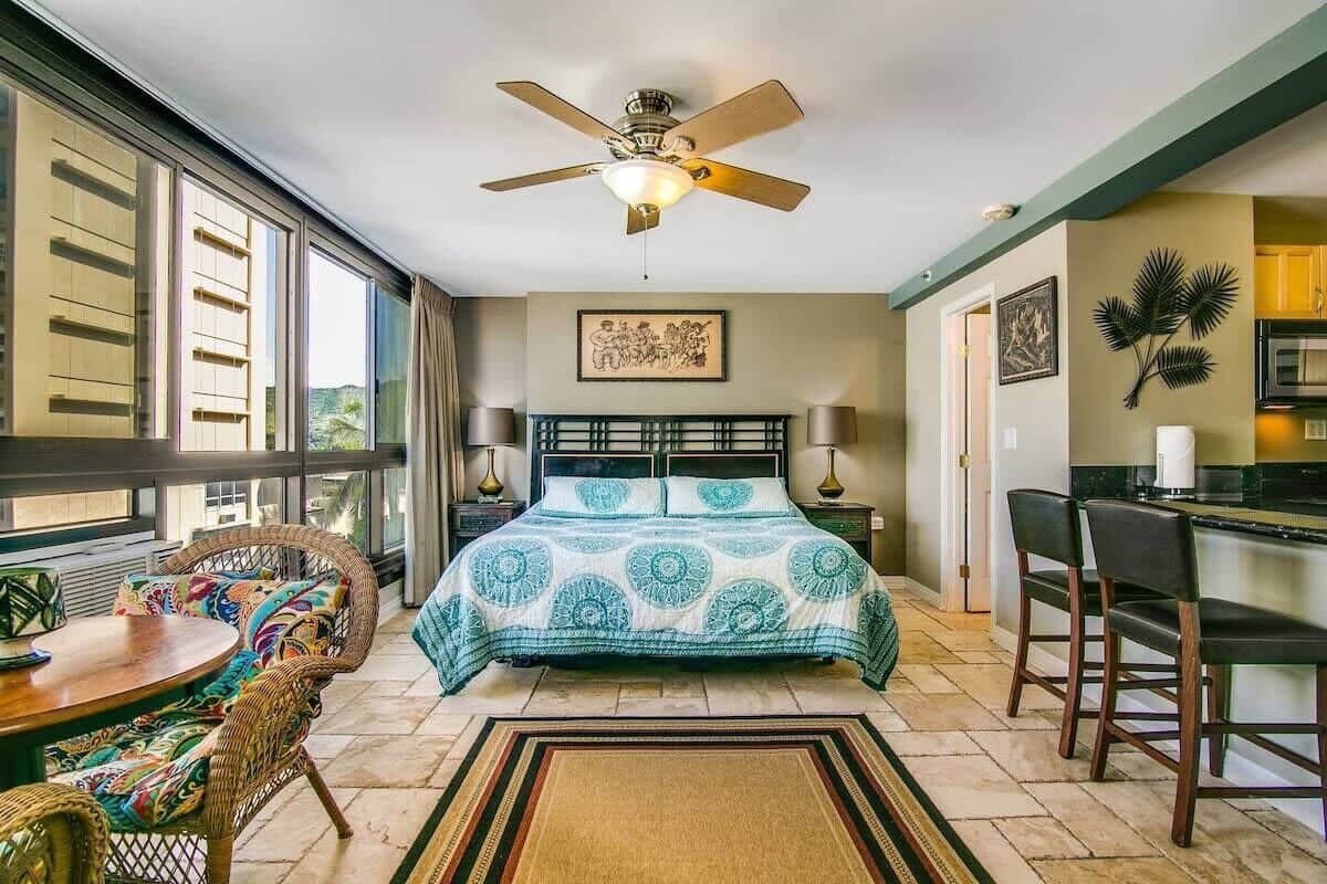 A bedroom with a bed, a ceiling fan, a dining table with chairs, and a view of the outdoors.