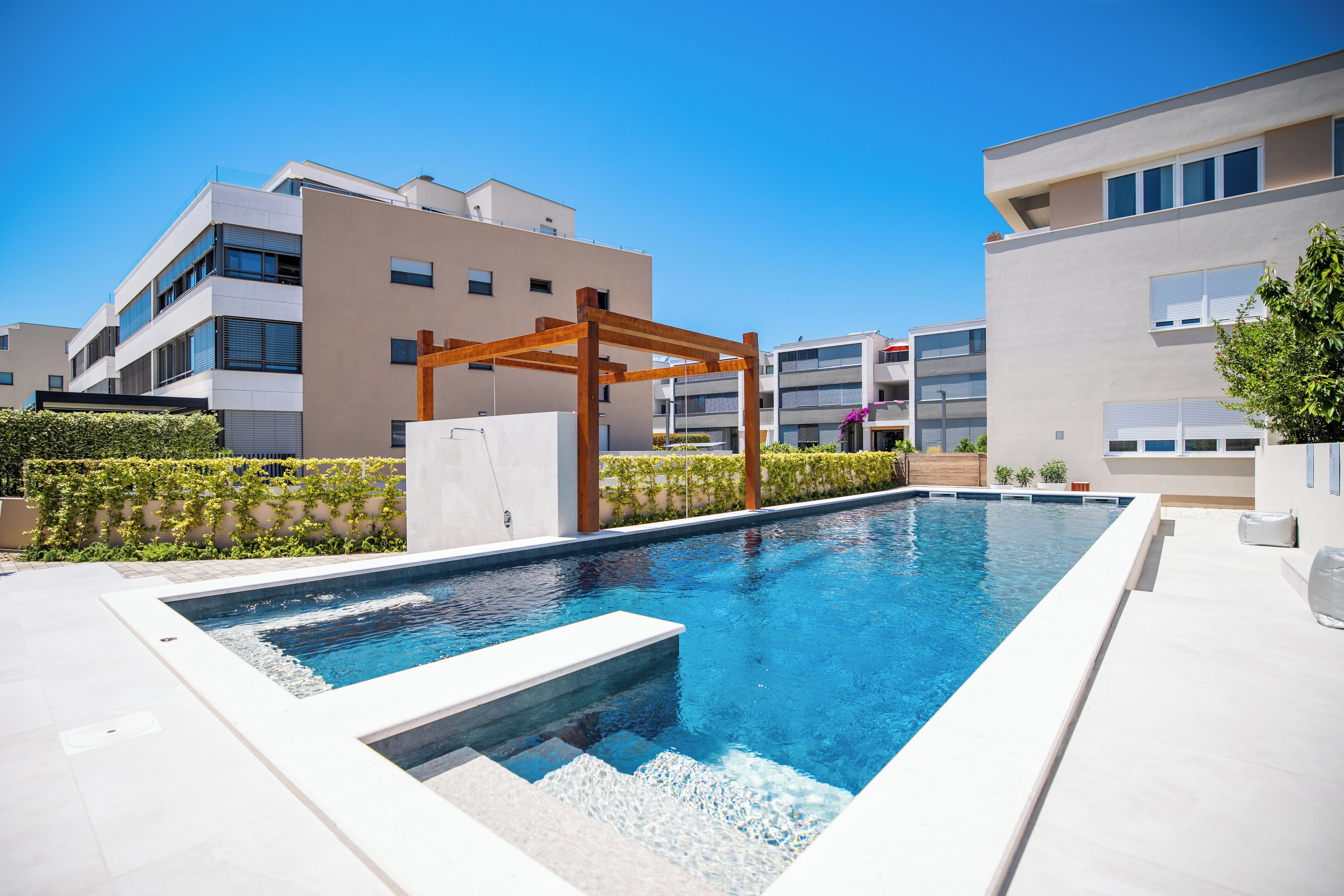 Una piscina moderna al aire libre con una pérgola de madera, rodeada por edificios contemporáneos y un seto bien cuidado.