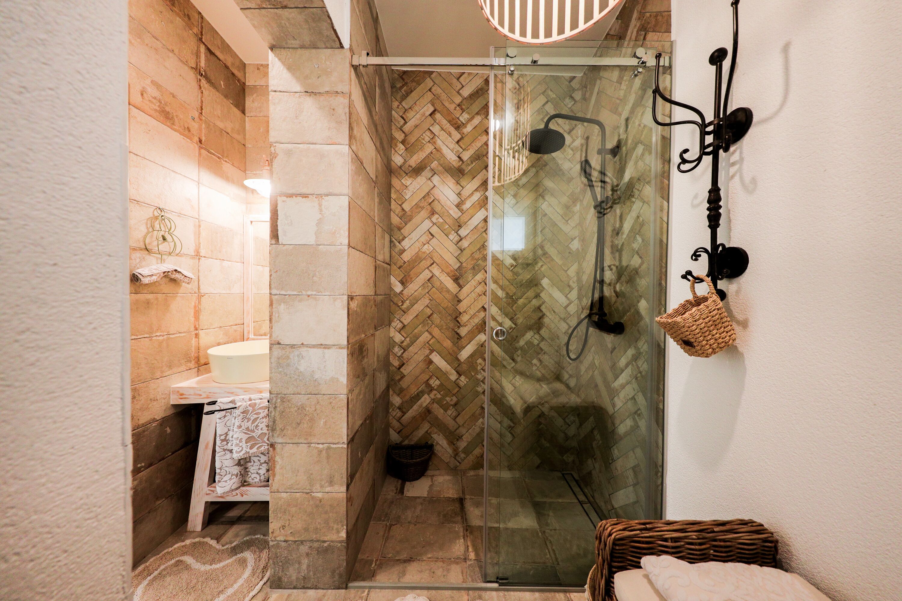 A modern bathroom with a herringbone tile pattern, a glass-enclosed shower, a sink with a white countertop, and a wicker chair.