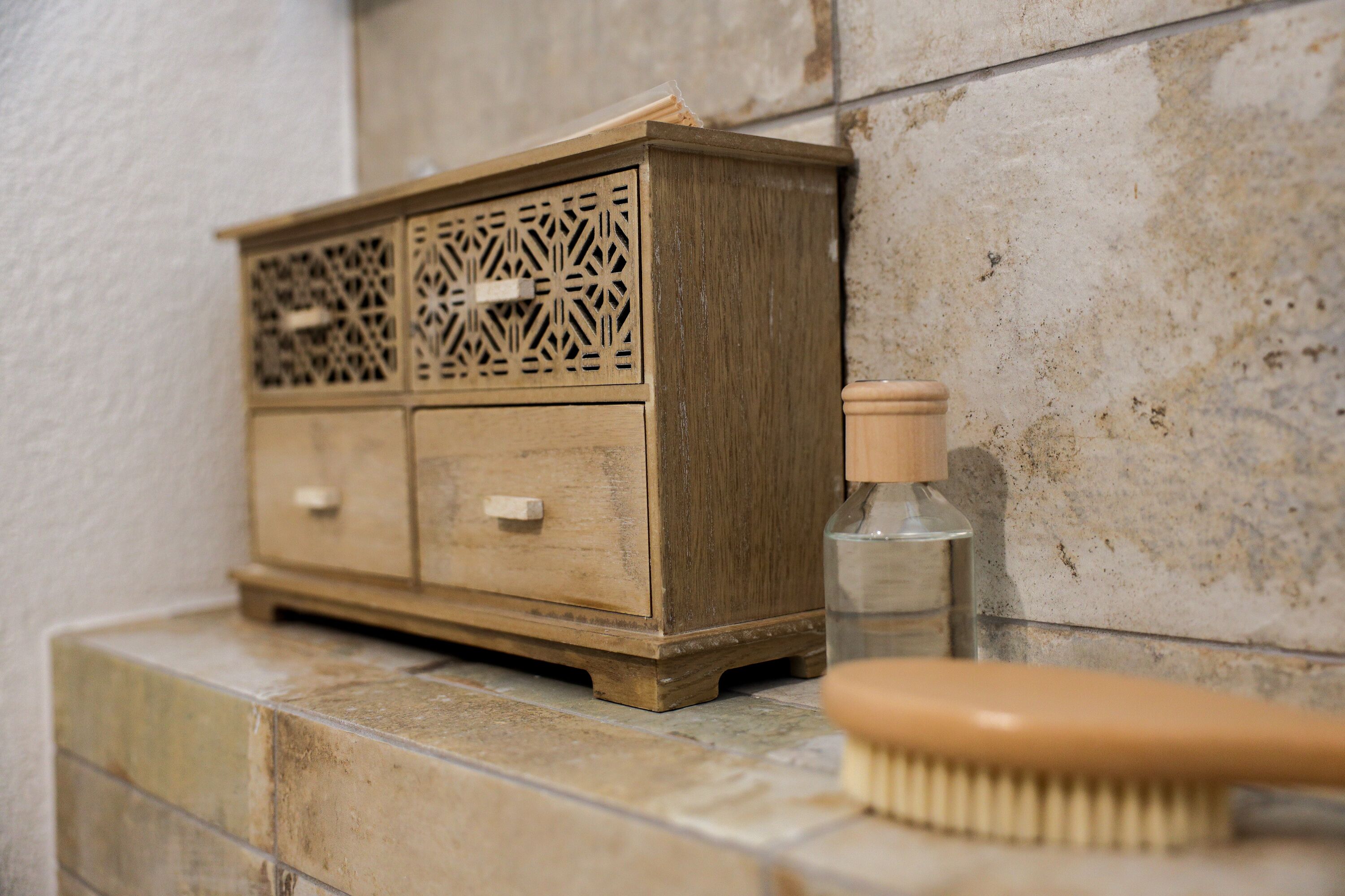 Un mueble de madera con diseños tallados elaborados, una botella pequeña y un cepillo de madera sobre una superficie de baldosas.