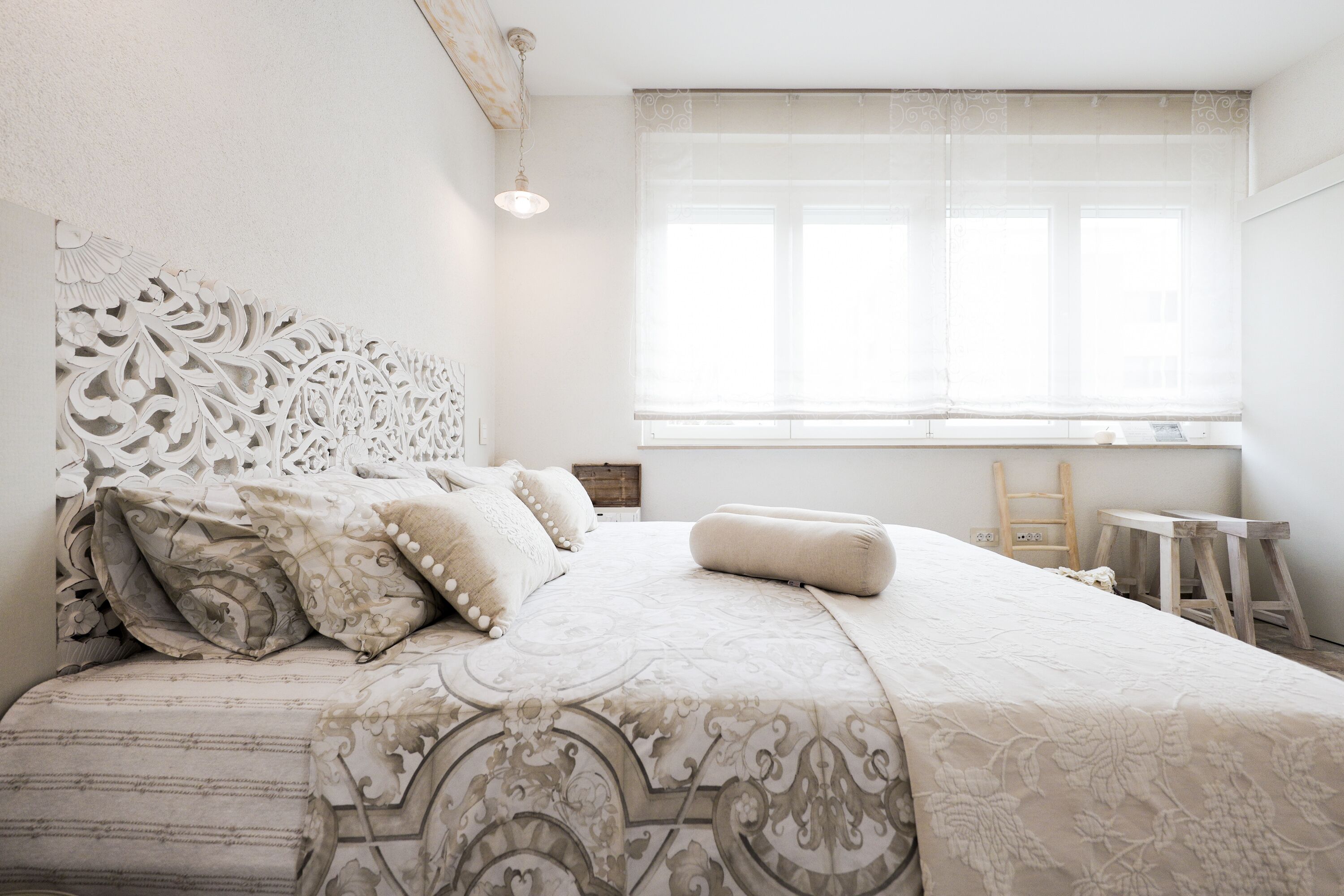 Una cama bien hecha con una colcha y almohadas estampadas, una ventana con cortinas de encaje y una escalera de madera apoyada en la pared.