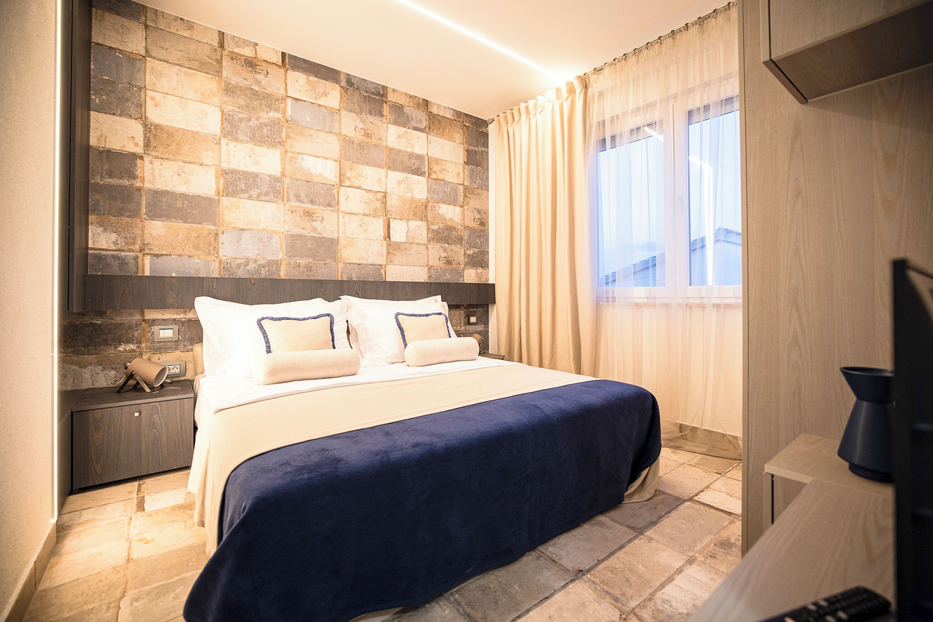A hotel room with a large bed, a nightstand, a television, and a stone-patterned wall.