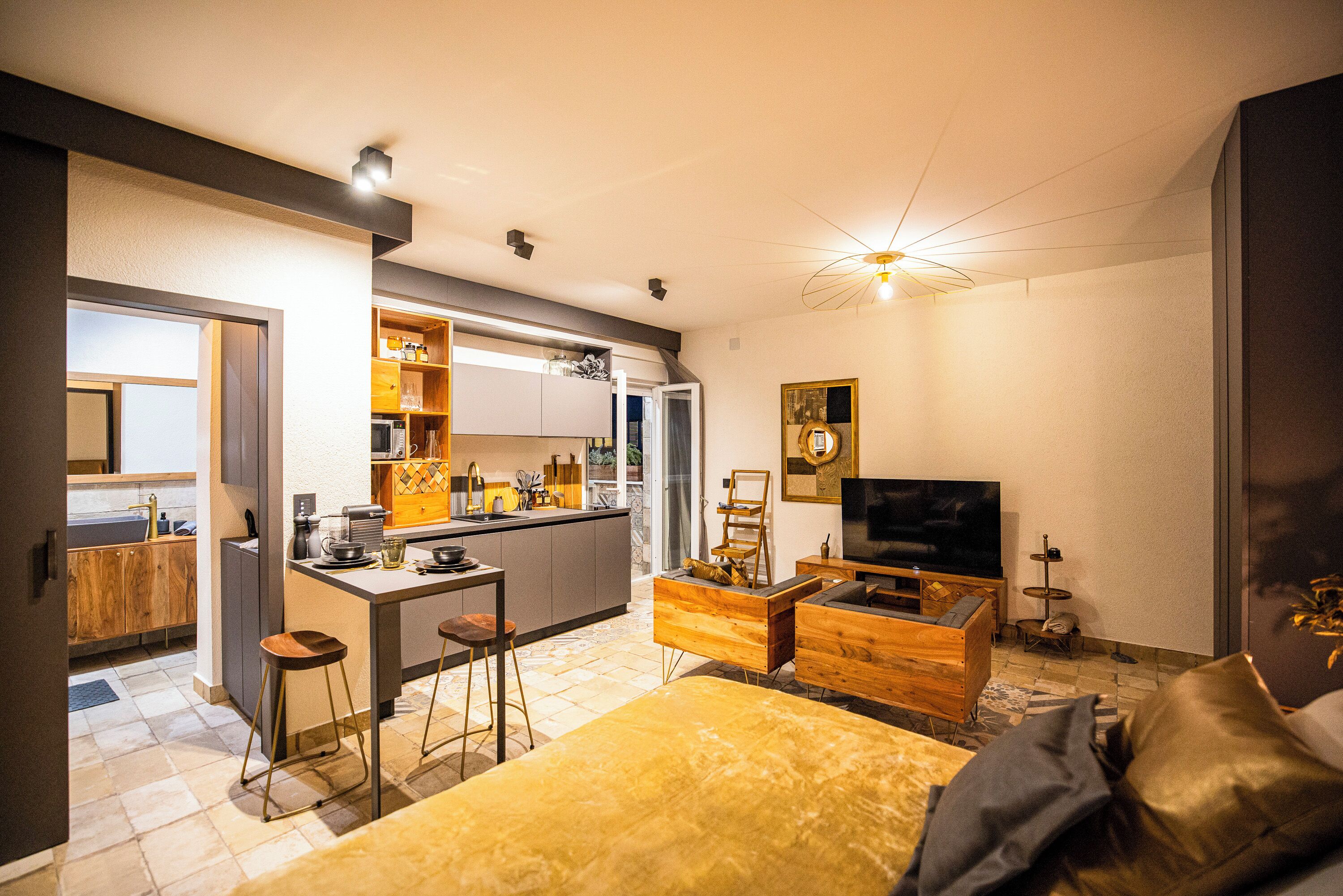 Una cocina moderna con una isla central, un área de comedor con sofá y televisor, y una cama grande con detalles dorados.