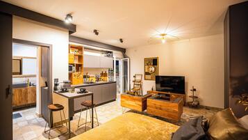 Una cocina moderna con una isla central, un área de comedor con sofá y televisor, y una cama grande con detalles dorados.