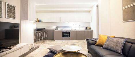 Una cocina moderna con armarios blancos, un comedor con un sofá negro y una mesa de centro redonda, y un televisor en la sala de estar.