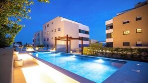 A swimming pool with a wooden pergola, surrounded by modern buildings and well-lit pathways. -  Ika Boutique Luxury Apartments (Zadar)