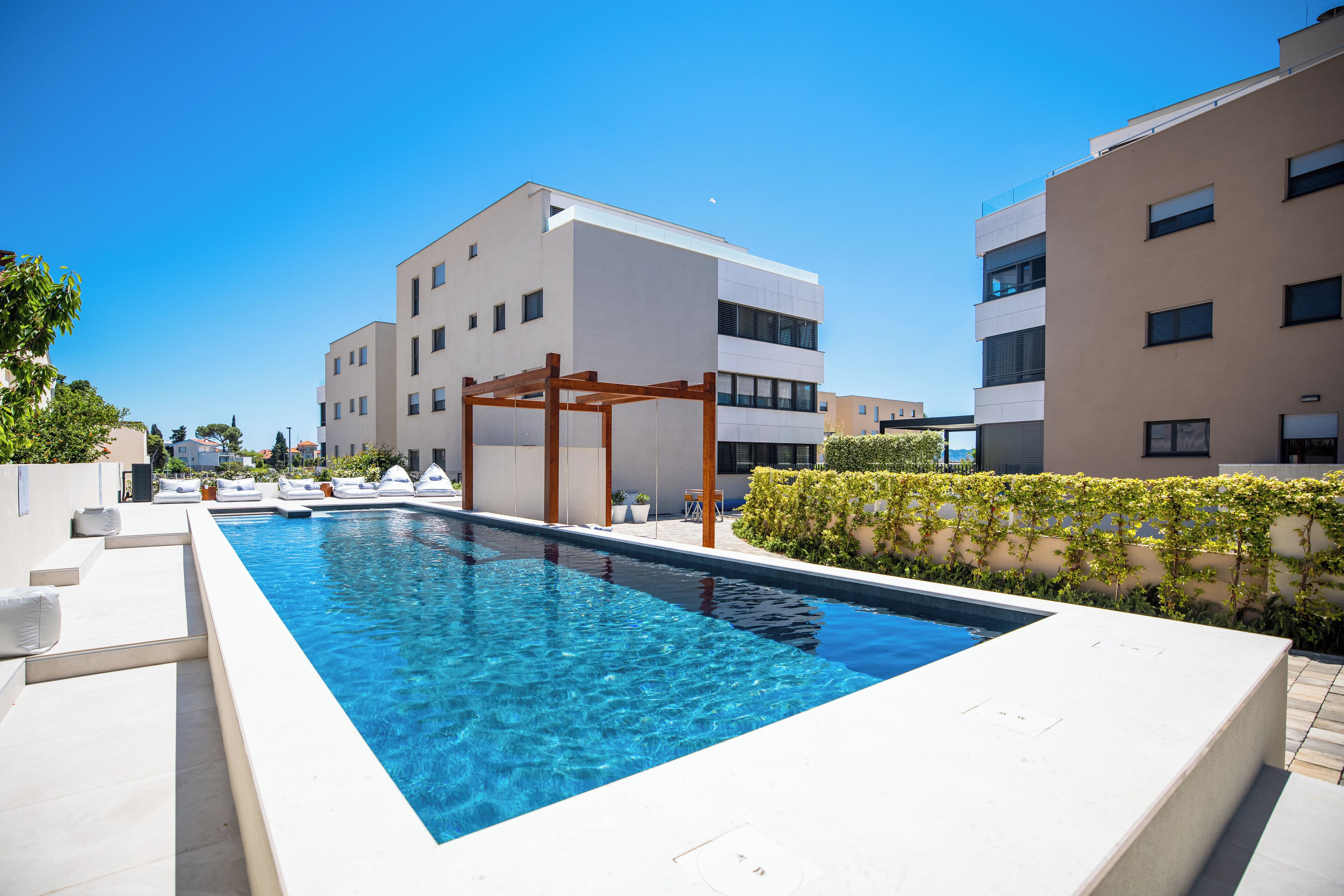 Un área de piscina moderna con piscina azul cristalina, pérgola de madera y terraza con baldosas blancas.