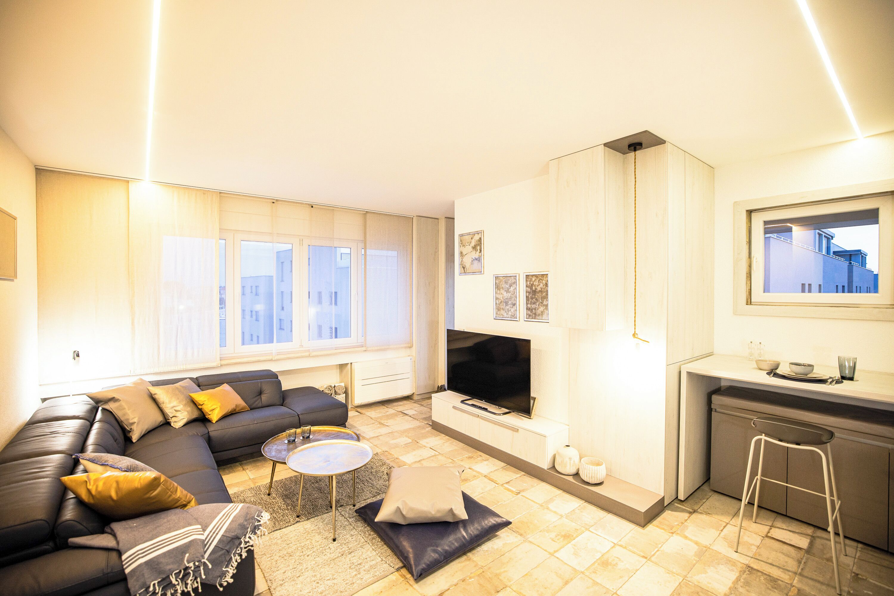 A modern living room with a sofa, coffee table, and TV. There is a kitchen area with a bar stool and a window overlooking the city.