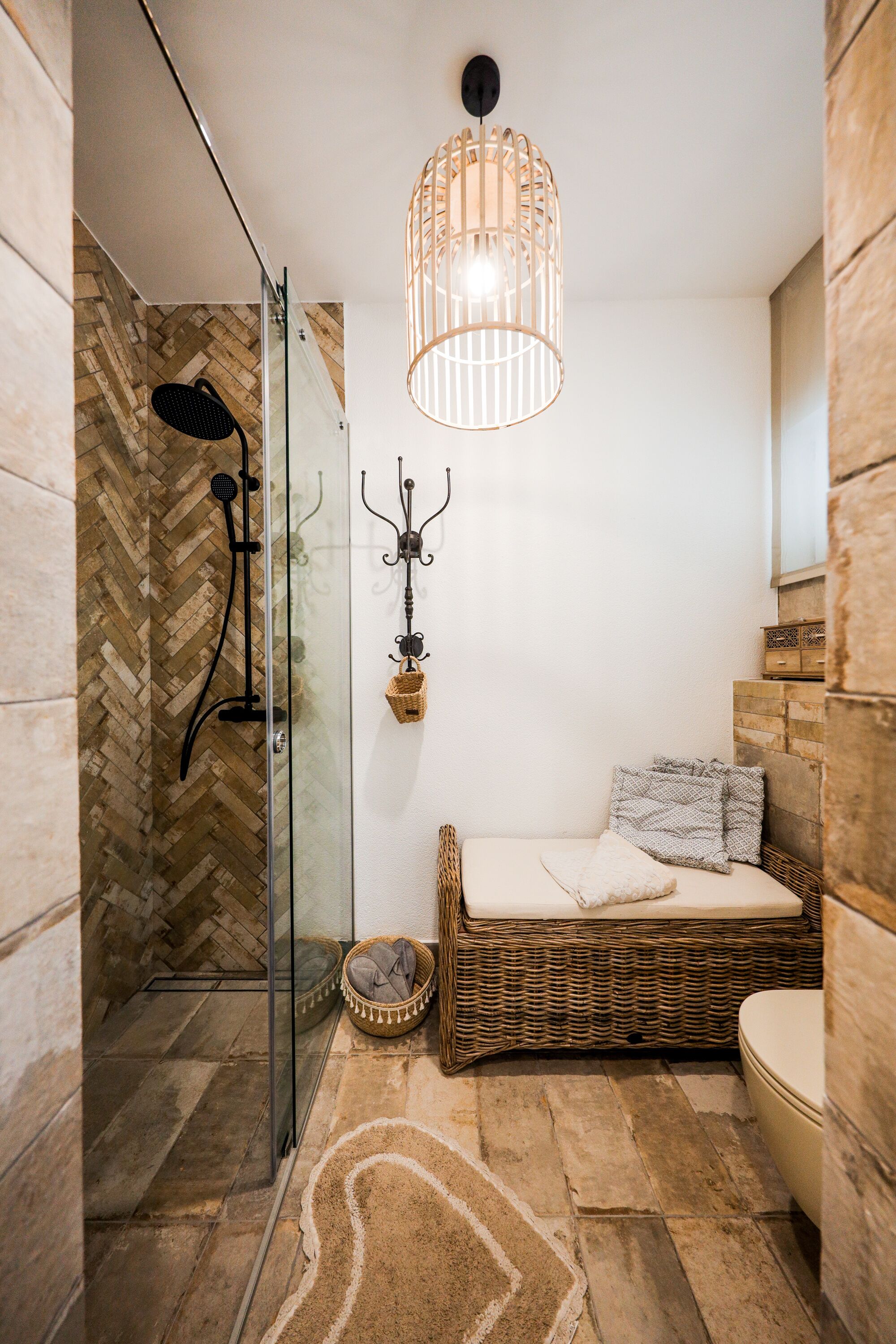 A bathroom with a glass shower enclosure, a wicker sofa with cushions, a hanging light fixture, and a small wicker basket on the floor.