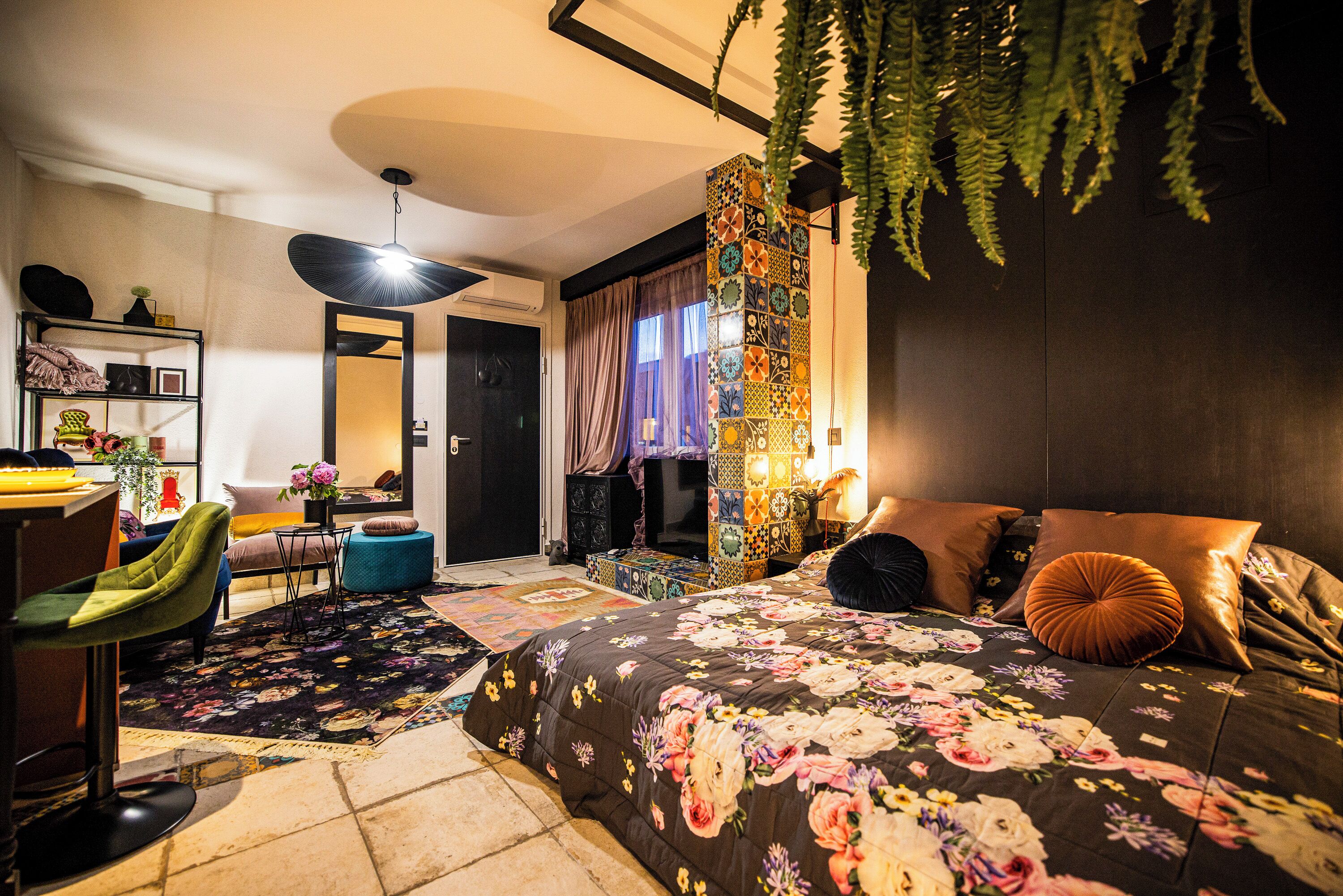A bedroom with a floral bedspread, a green chair, a desk with a lamp, and a hanging plant.