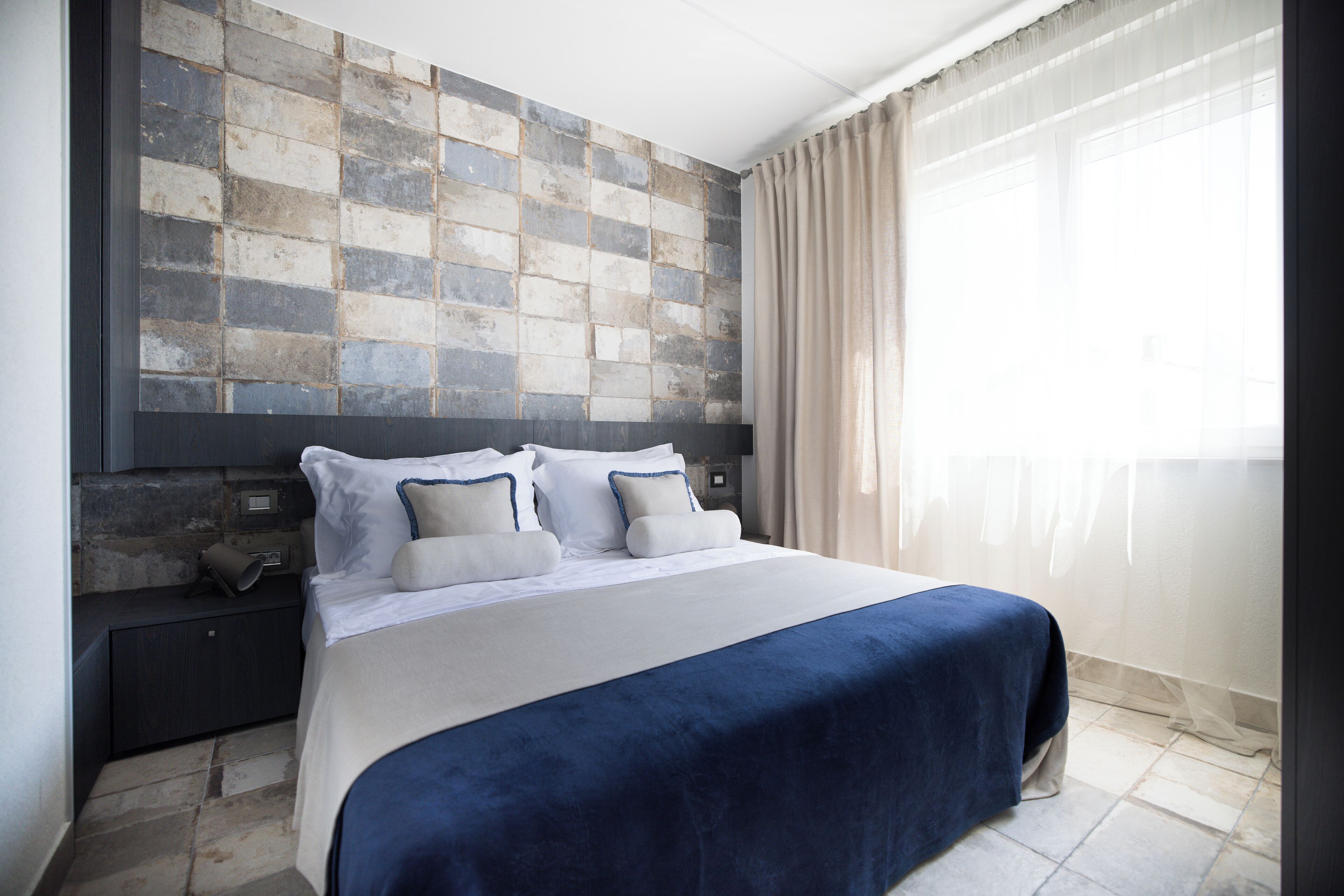 Een moderne hotelkamer met een groot bed, een hoofdbord met een structuurwand, een nachtkastje en een raam met gordijnen.