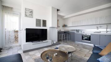 Una sala de estar moderna con un televisor de pantalla plana, dos mesas auxiliares, un sofá y una zona de cocina con taburetes de bar.