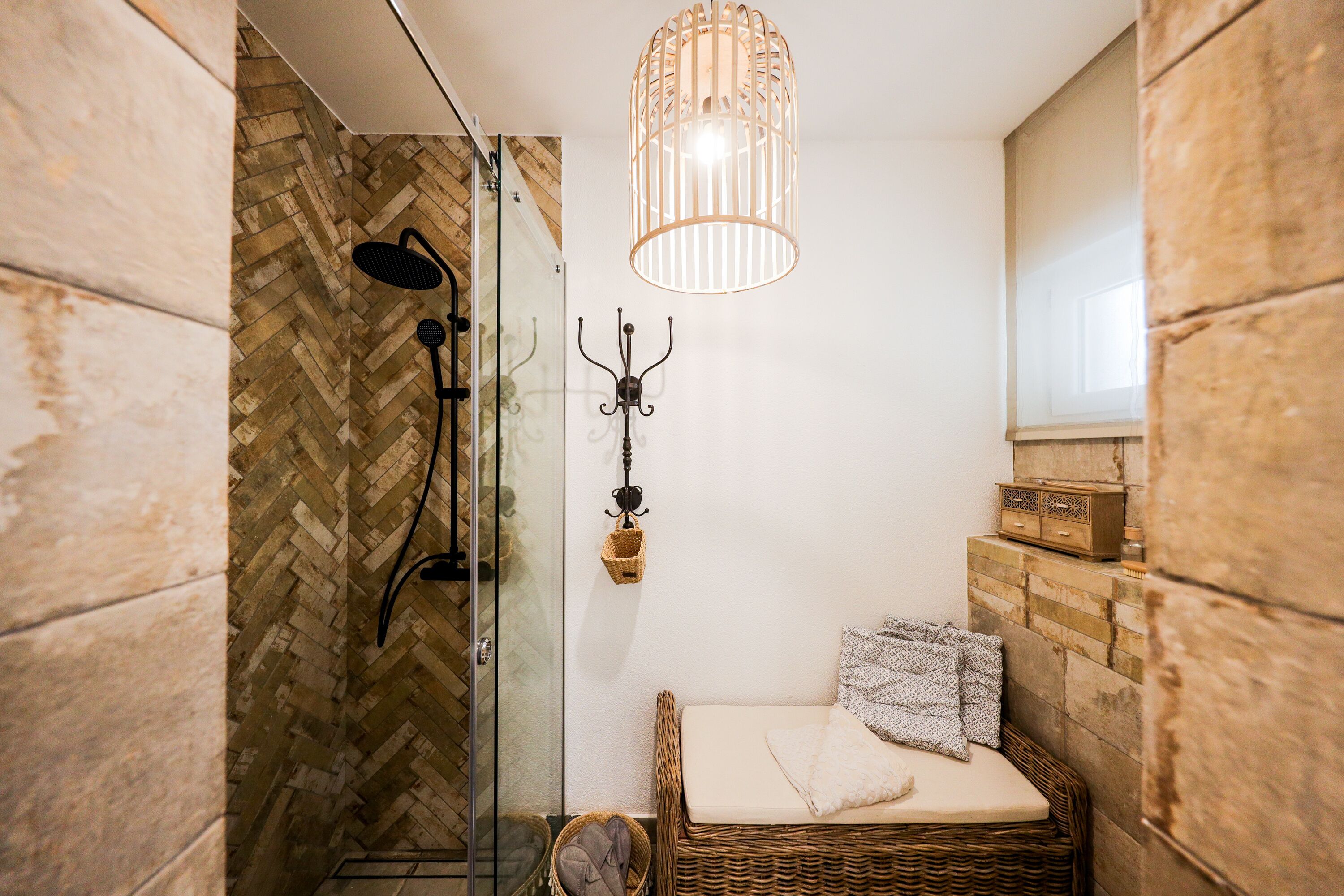 A bathroom with a glass shower enclosure, a wicker bench with cushions, a hanging light fixture, and a wooden wall.