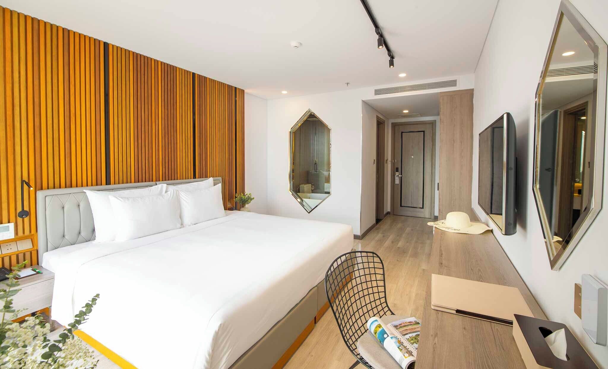 A modern hotel room with a large bed, a desk with a chair, a mirror, and a television.