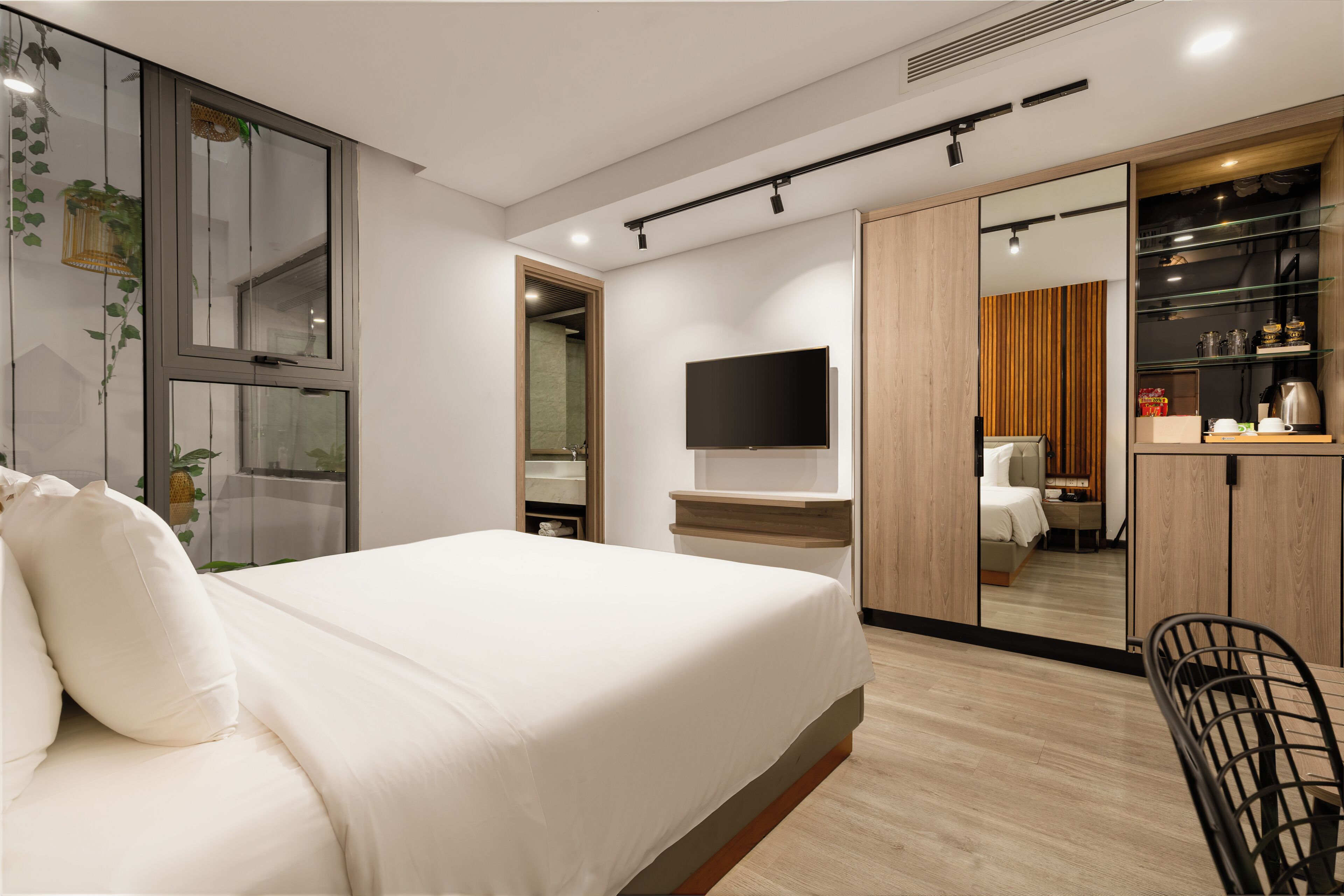 A modern hotel room with a large bed, a flat-screen TV, a wooden wardrobe, and a small kitchenette.