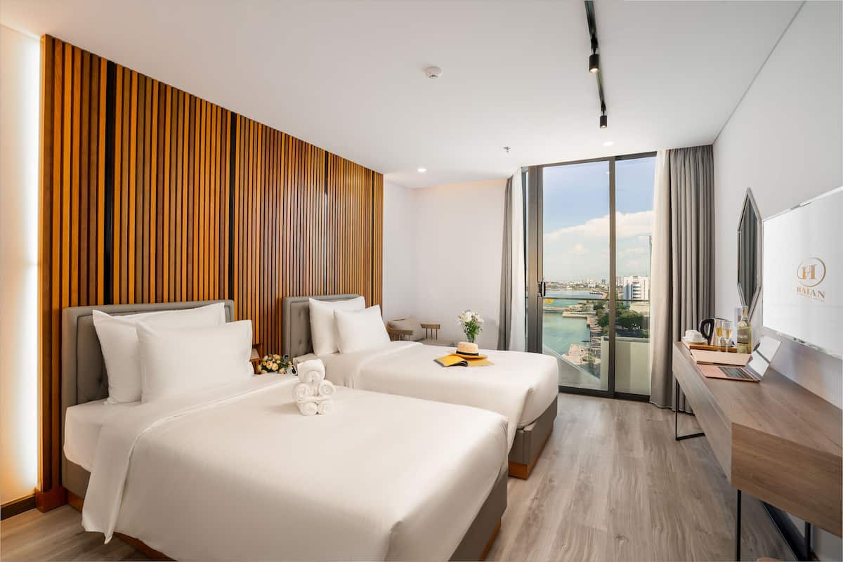 Quarto de hotel com duas camas, uma escrivaninha e uma janela grande com vista para a cidade.