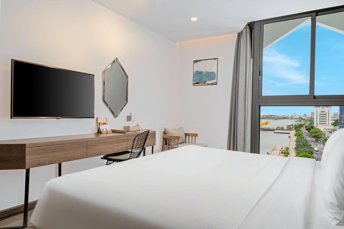 Quarto de hotel moderno com uma cama grande, uma escrivaninha com cadeira, televisão de tela plana e vista para a cidade através de amplas janelas.
