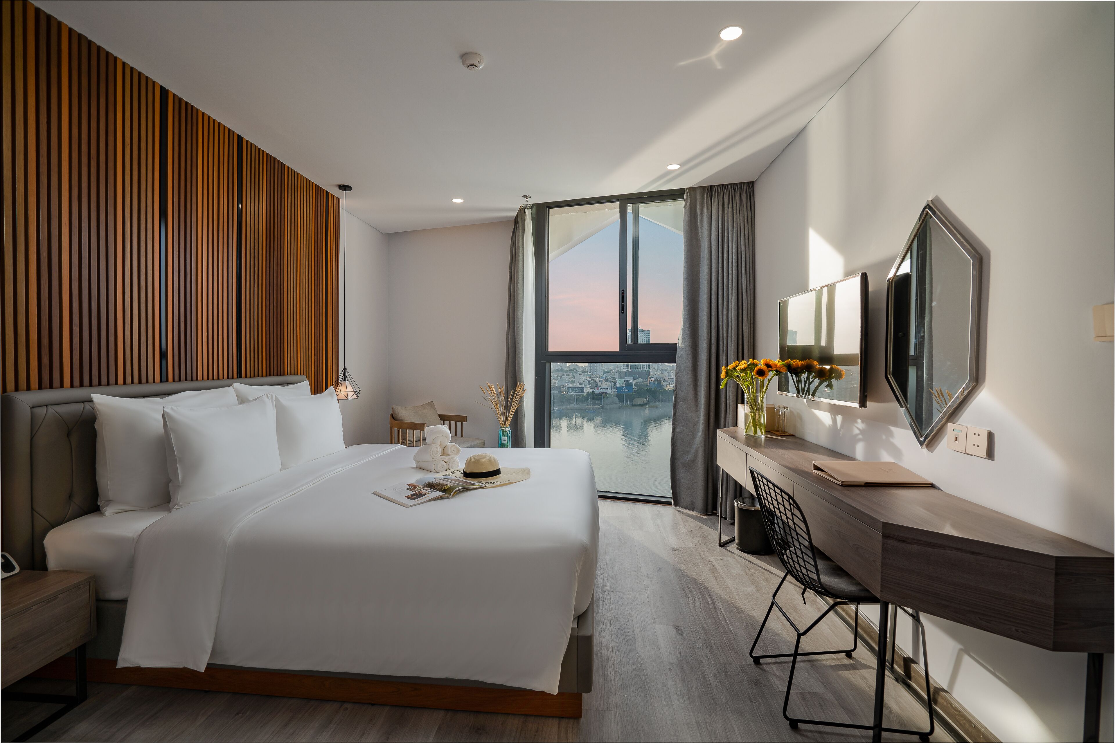 A modern hotel room with a large bed, a desk, and a view of a cityscape through a large window.