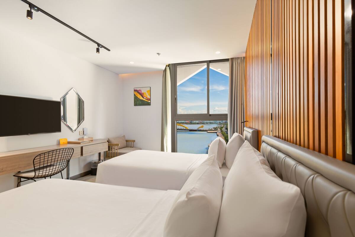 Quarto de hotel moderno com duas camas, uma mesa de trabalho, uma cadeira, televisão e uma janela ampla com vista para a água.