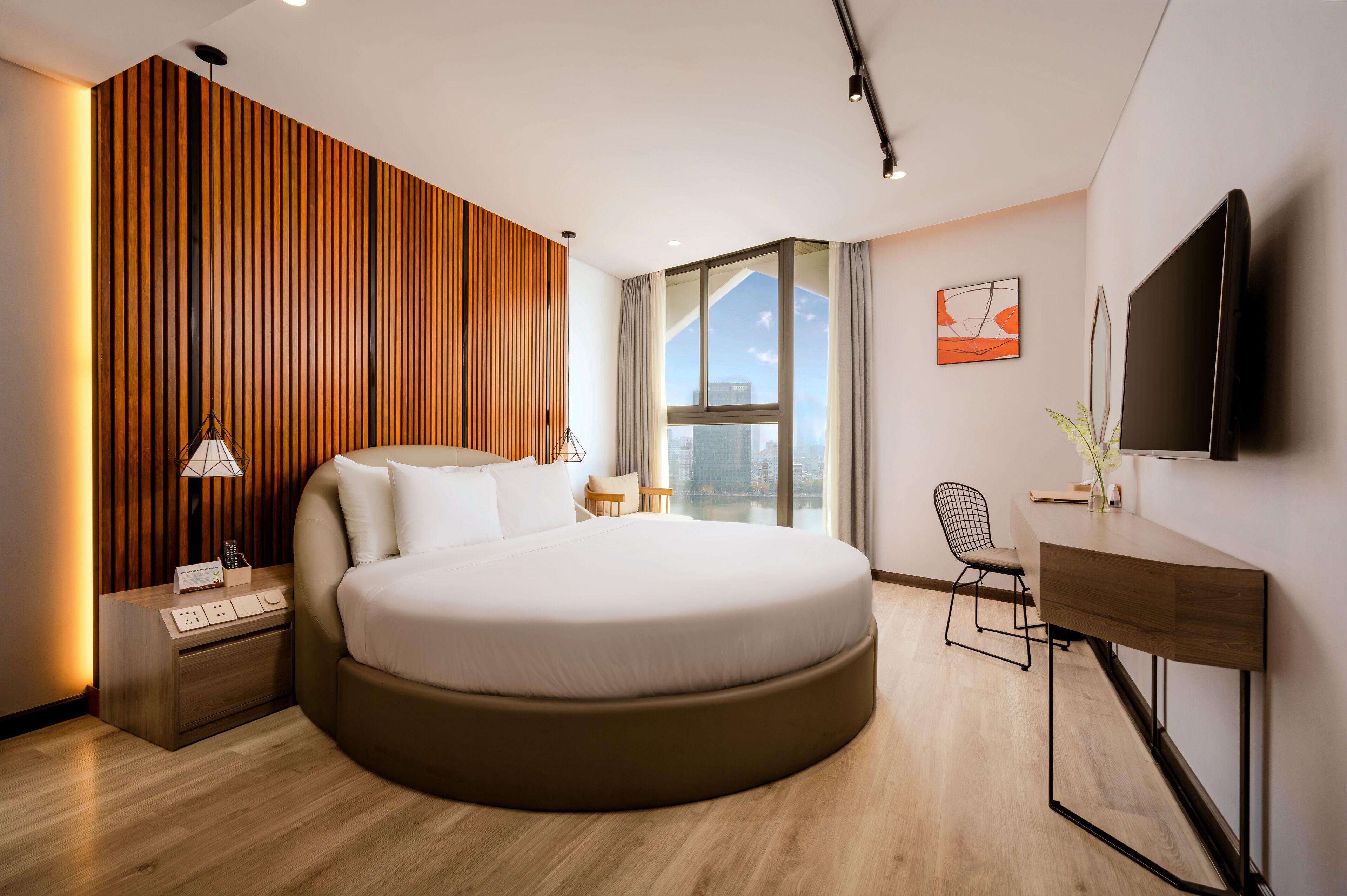 A modern bedroom with a large bed, a desk, a TV, and a city view through the window.