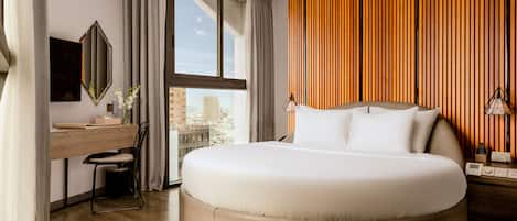 A modern hotel room with a large bed, a desk with a chair, a mirror, and a view of the city.