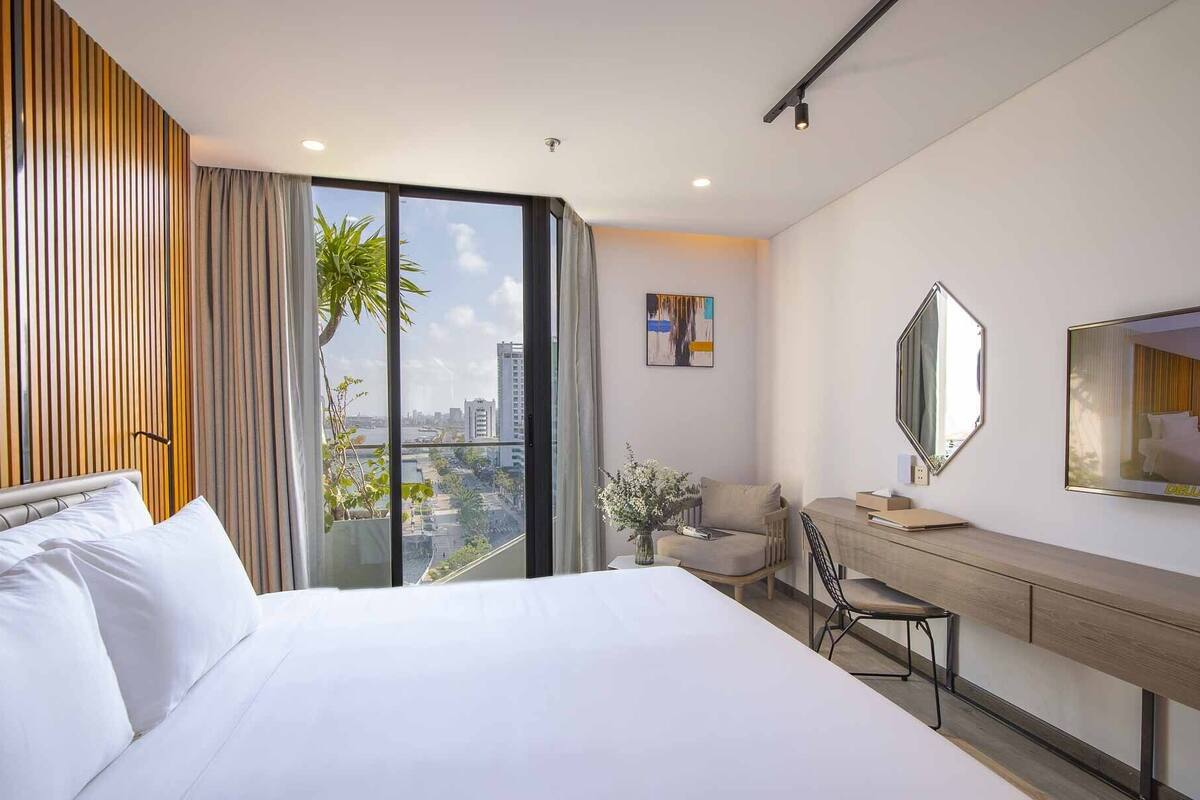 Quarto de hotel moderno com uma cama grande, uma escrivaninha, uma cadeira, um espelho e vista para a cidade.