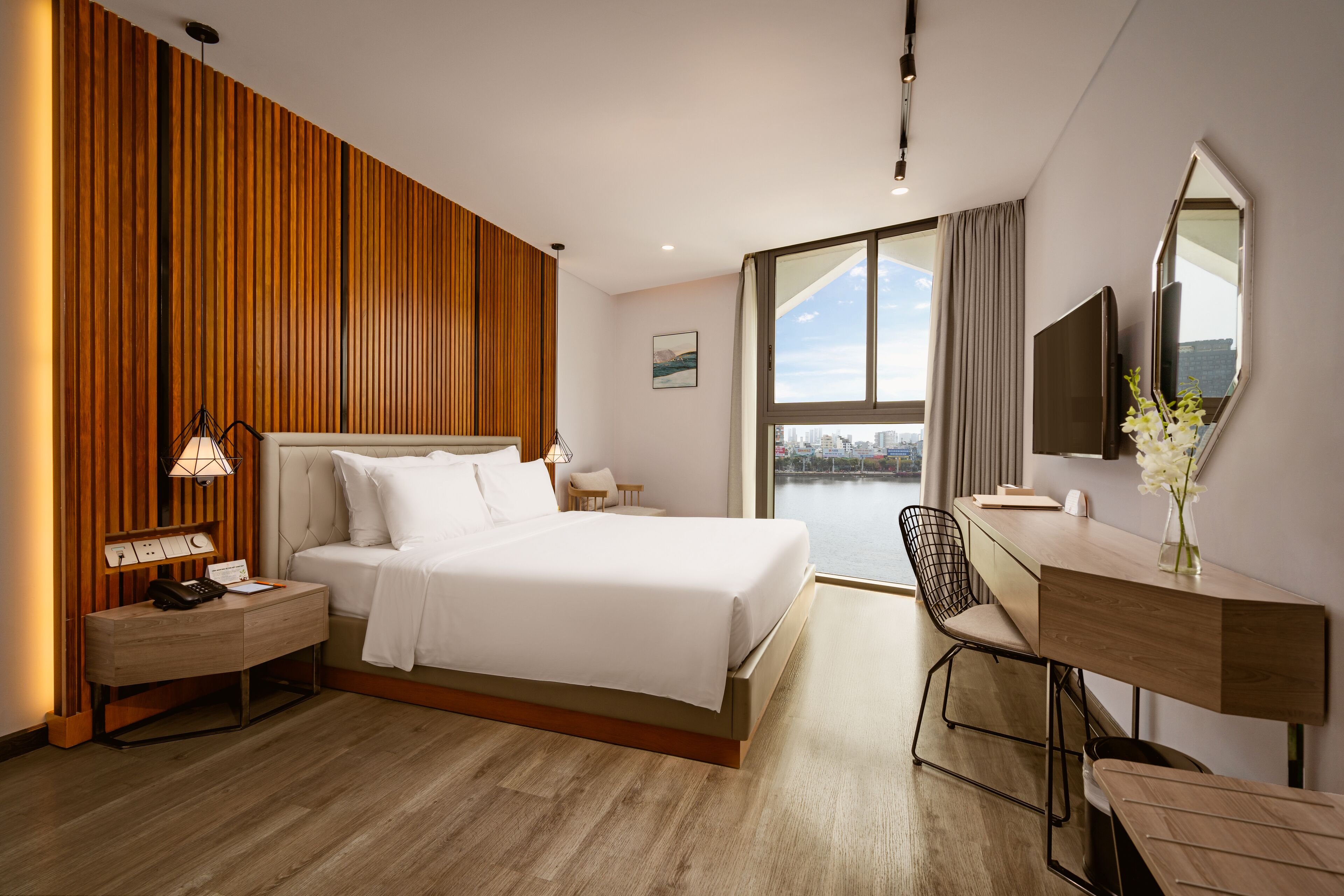 A modern hotel room with a large bed, a desk with a chair, a flat-screen TV, and a view of the city through large windows.