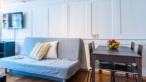 A modern living room with a grey sofa, a wooden dining table with chairs, a TV mounted on the wall, and a fruit bowl on the table. - ZH Niederdorf II - Hitrental Apartment (Zürich)