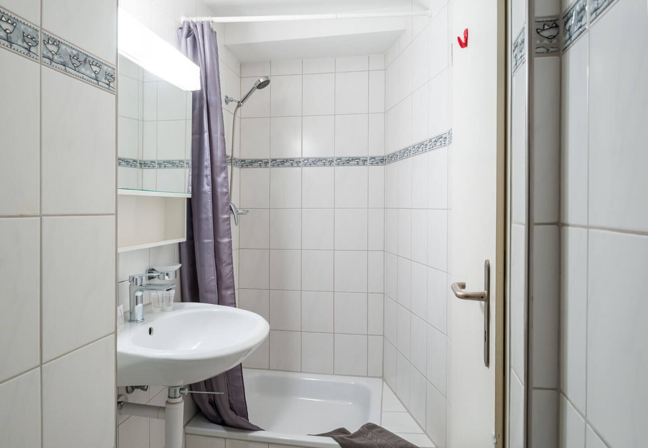 Une salle de bain avec un lavabo blanc, une douche avec un rideau violet et des murs carrelés.