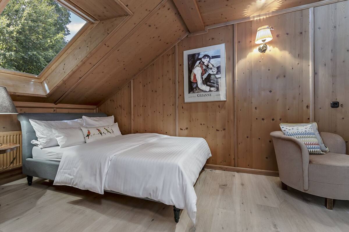 Une pièce confortable avec un lit, une chaise, des murs en bois, une lucarne et un tableau encadré.