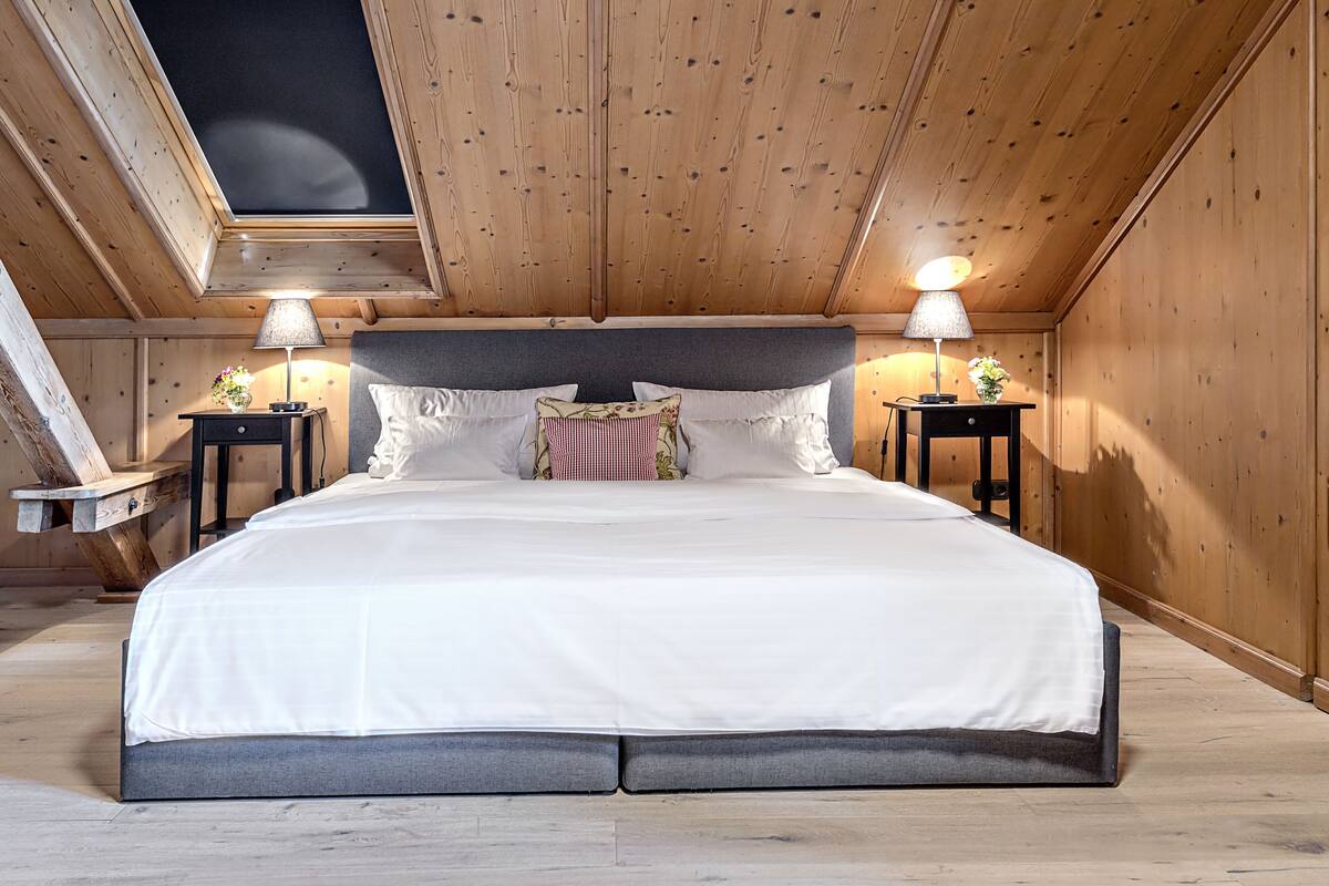 Un grand lit avec une tête de lit grise, dans une chambre en bois dotée d’une lucarne. On y trouve deux tables de chevet avec des lampes.