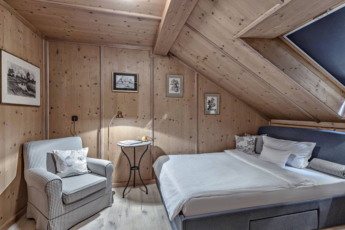 Une pièce chaleureuse aux murs en bois, avec un fauteuil à carreaux, une petite table et un lit recouvert de draps blancs.