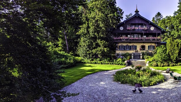 Eine zweistöckige Chalet mit Balkon, umgeben von üppigem Grün und einem Kiesweg zum Eingang.