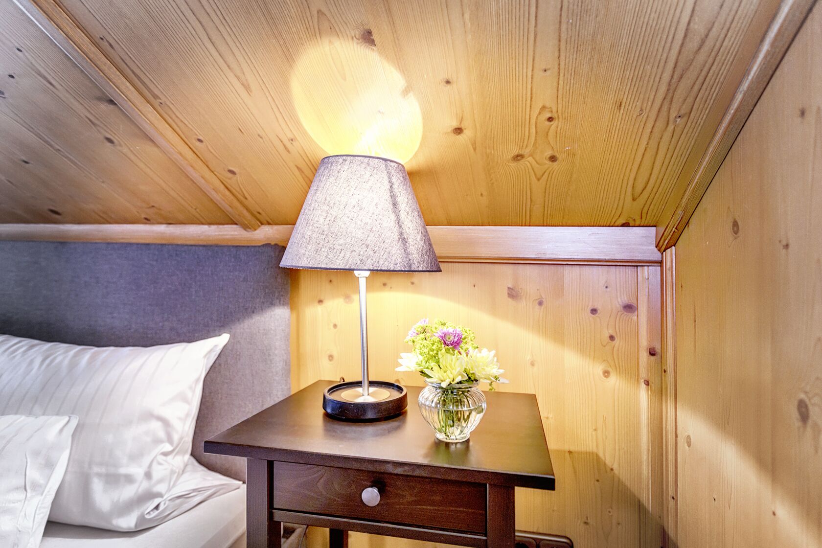 A wooden cabin room with a bed, a nightstand with a lamp, and a vase with flowers.