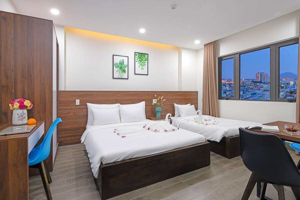 Quarto de hotel com duas camas, uma escrivaninha, uma cadeira, um vaso com flores e vista para a cidade.