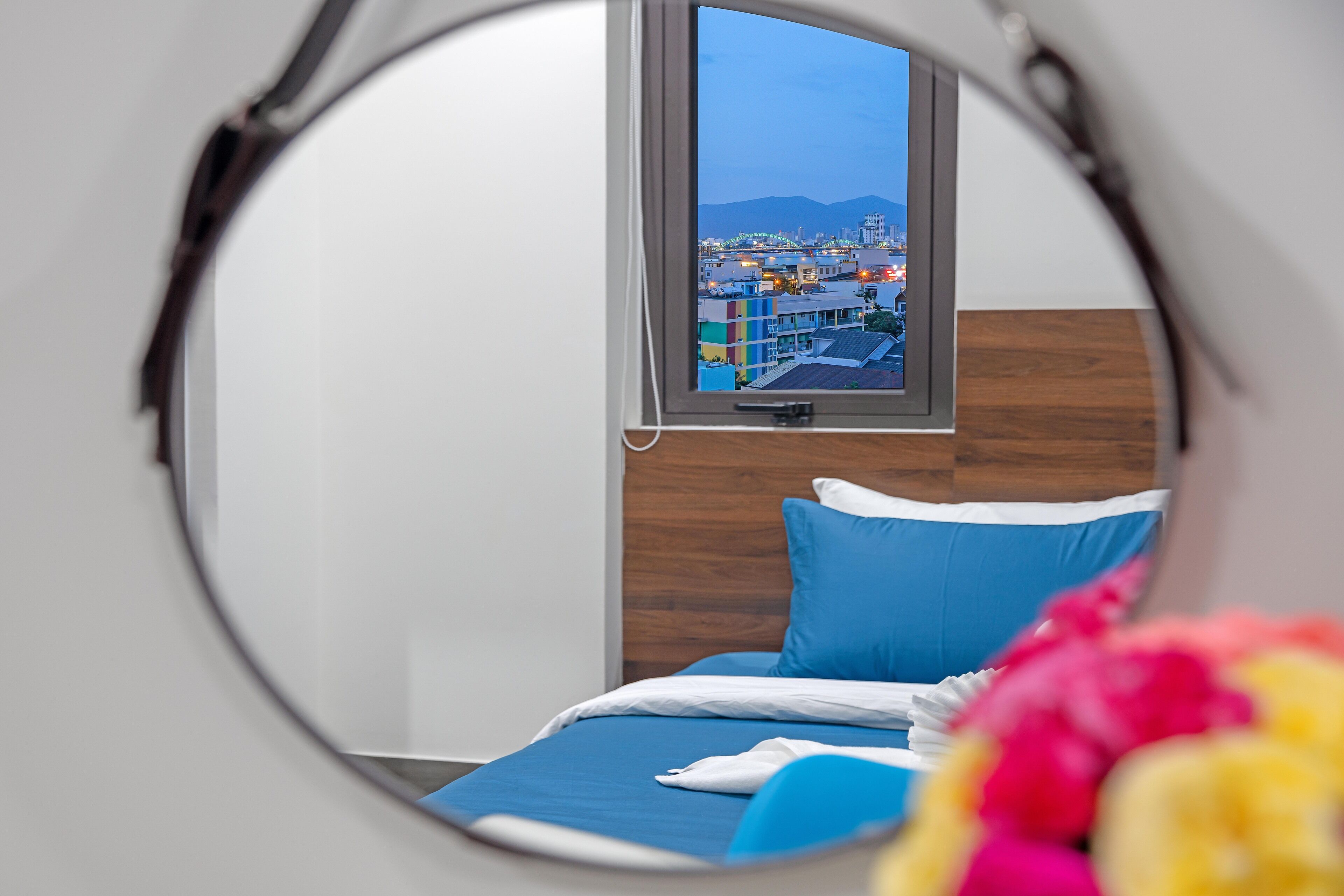A bedroom with a circular window, a bed with blue bedding, and a view of a cityscape.