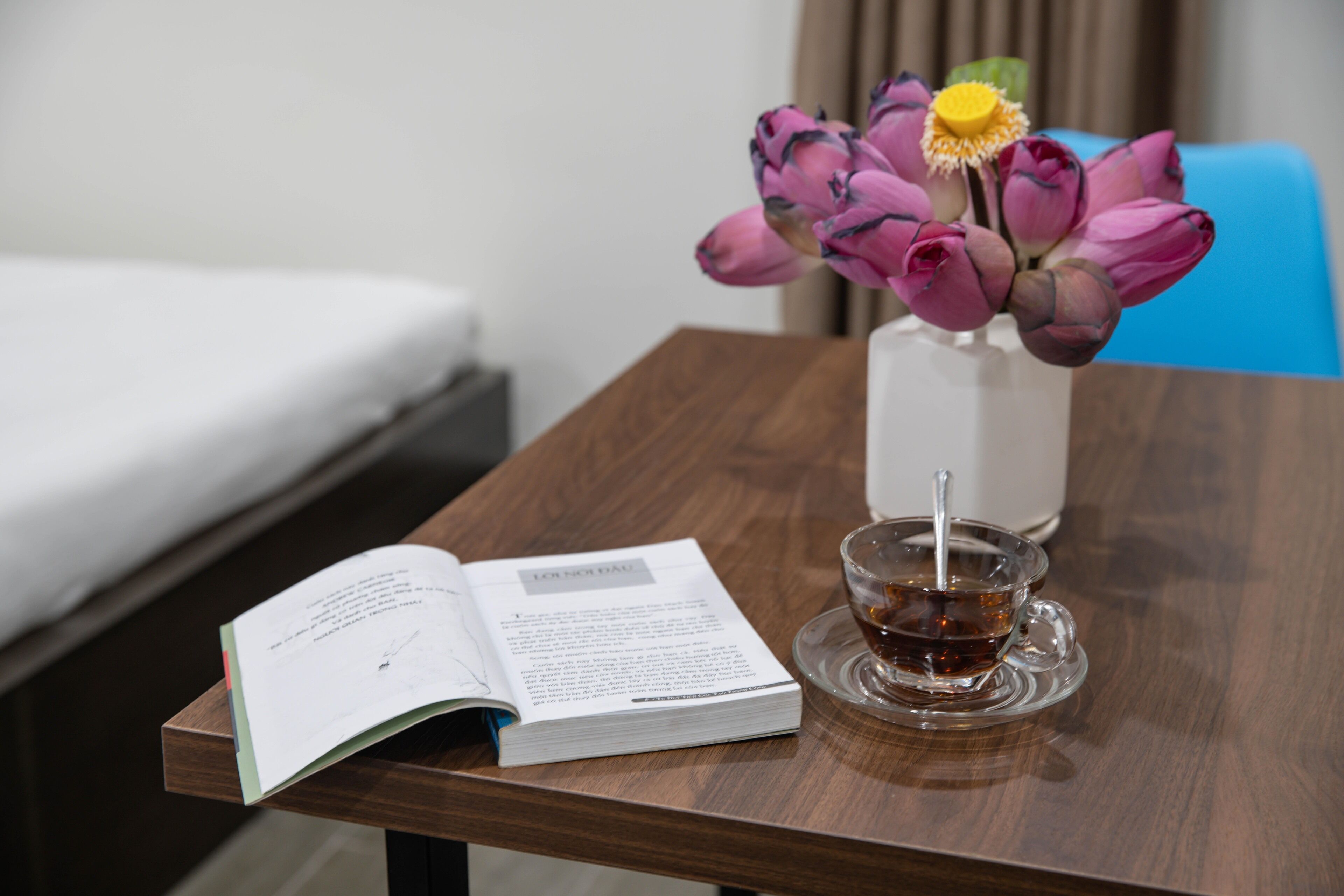 A wooden table with a book, a glass cup with a tea spoon, and a vase with pink flowers.