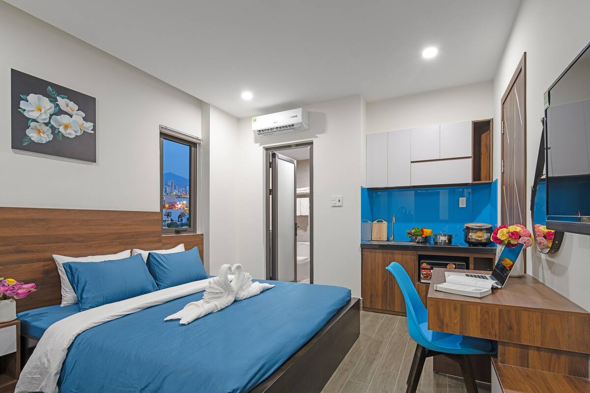 Quarto de hotel com uma cama, escrivaninha, cadeira, janela e um quadro na parede.