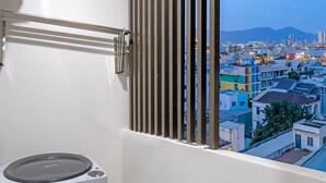Un balcón con lavadora, una ventana con persianas y vista a la ciudad.