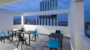 Una terraza al aire libre con mesas y sillas, vista a un paisaje urbano y un edificio moderno al fondo.
