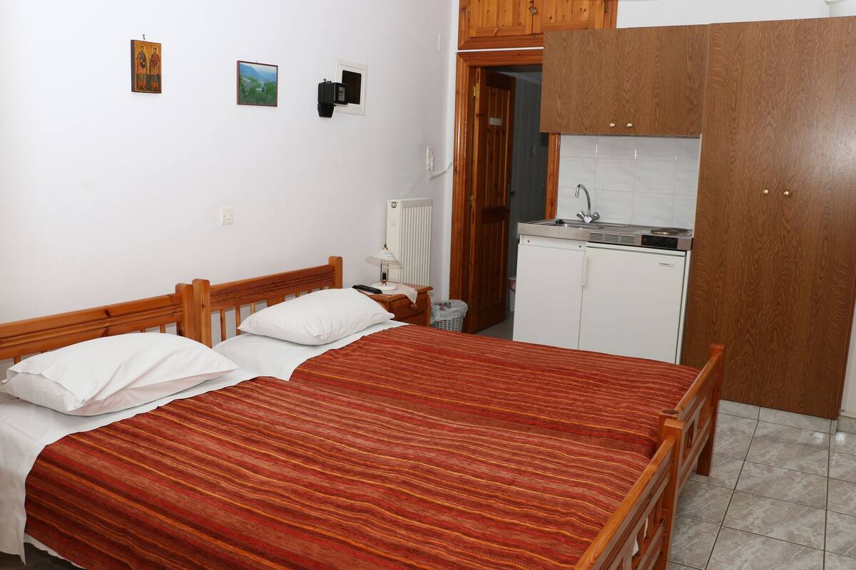 Un dormitorio con cama de madera, almohadas blancas, colcha a rayas rojas, mesita de noche, lámpara, radiador, y una pequeña cocina con lavabo y estufa, además de armarios de madera.
