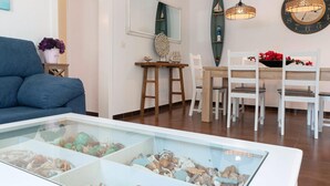 Una sala de estar moderna con un sofá azul, una vitrina de vidrio con objetos decorativos, una zona de comedor con sillas blancas y una mesa de madera, y un reloj de pared.