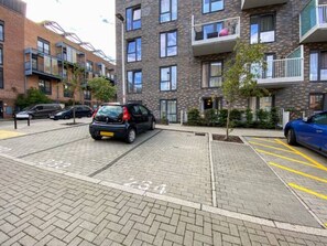 Ein Parkplatz mit markierten Stellplätzen, ein mehrgeschossiges Gebäude mit Balkonen und ein Baum im Vordergrund.