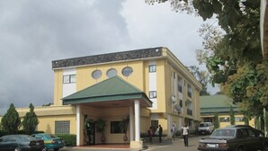 A building with a yellow facade, a green roof, and a covered entrance. There are cars parked in front and people walking around. - Lamonde Guest House (Owerri)